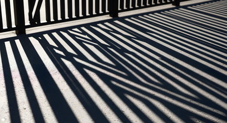 An abstract, high-contrast image showing the dark shadows of a fence or railing cast onto a light-colored, textured ground. The repeating lines and angles create a strong geometric and graphic pattern.の素材