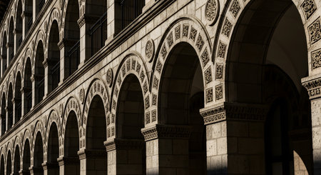 A low-angle perspective of a row of ornate, carved stone arches on the facade of a classical or Romanesque-style building. Strong sunlight creates deep shadows, highlighting the architectural details and textures. The image represents history, architecture, and timeless design.の素材