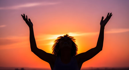 A silhouette of a joyful woman with curly afro hair, raising her arms in worship or celebration against a vibrant orange and purple sunset. Her head is tilted back in happiness. The image represents freedom, gratitude, and spirituality.の素材