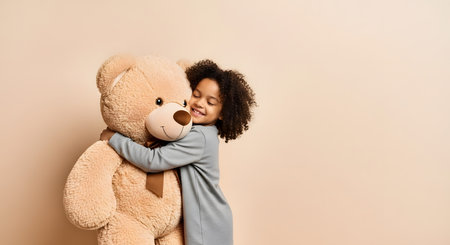 A happy young girl with curly hair and dark skin smiles with her eyes closed while hugging a large, soft, brown teddy bear. The child is standing against a plain beige background, with ample copy space to the right.の素材