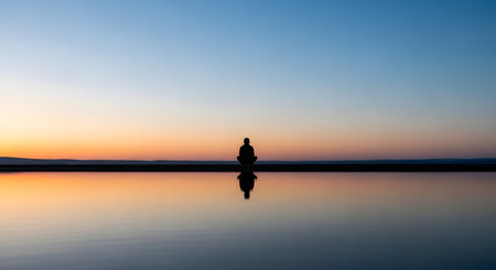 The silhouette of a person sitting in a meditation pose is perfectly reflected in still, calm water. The scene is set against a beautiful gradient sunset sky, with colors ranging from deep blue to warm orange. The image conveys peace, tranquility, mindfulness, and balance.の素材