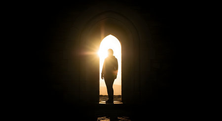 A silhouette of a person stands in the middle of a dark, stone arched doorway. Bright, warm sunlight streams through the arch from the outside, creating a stark contrast and a 'light at the end of the tunnel' effect. This image symbolizes hope, new beginnings, opportunity, or a spiritual journey.の素材