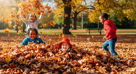 Four happy, diverse children are playing in a large pile of colorful autumn leaves in a sunny park. One girl throws leaves in the air, a boy peeks out from the pile with a red hat, and another boy runs towards them.の素材