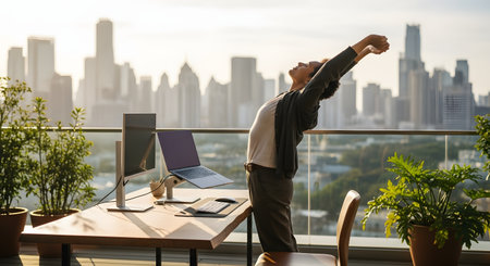 A person stretches their arms back while standing at an outdoor desk on a high-rise balcony. The standing desk has a laptop and monitor, overlooking a hazy city skyline at sunrise or sunset.の素材
