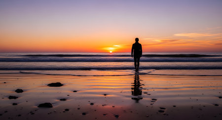 A person in a dark shirt, seen as a silhouette, stands on a wet sandy beach, looking at the sunset. The sun is on the horizon, casting a vibrant orange glow over the sky and reflecting on the ocean waves and wet sand.の素材