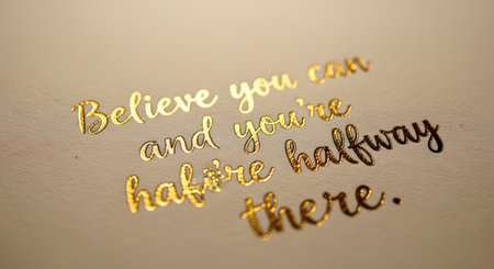A close-up, macro shot of an inspirational quote: 'Believe you can and you're halfway there.' The text is written in an elegant, shiny gold foil script on a textured, off-white paper. The shallow depth of field highlights the texture and shine.の素材