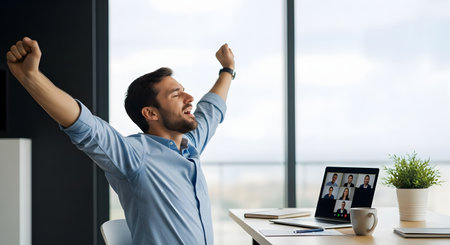 A joyful businessman stretches his arms up in victory while sitting at his desk in front of a laptop during a video call. He looks relieved and excited, celebrating a success or achievement with colleagues online.の素材