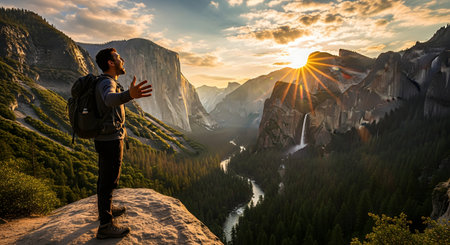 A male hiker with a backpack stands on a cliff at Tunnel View in Yosemite National Park, his arms outstretched in awe. He overlooks the iconic valley, with El Capitan and Bridalveil Fall visible, as a dramatic sunburst sets behind the mountains.の素材