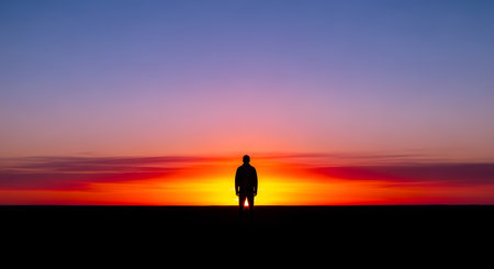 A solitary silhouette of a person stands against a vibrant and colorful sunset sky, gazing at the horizon. The minimalist composition highlights the contrast between the dark figure and the gradient of orange and purple light.の素材