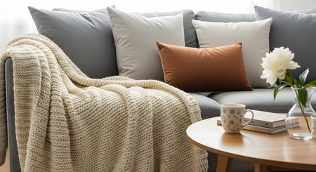 A cozy living room detail features a grey sofa with a chunky knit blanket and pillows. A wooden coffee table in the foreground holds a cup of coffee, a book, and a vase with a flower, creating a relaxing atmosphere.の素材