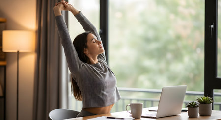 A young woman stretches her arms overhead with eyes closed while sitting at her desk in a home office. She is taking a relaxing break from working on her laptop to relieve stress and improve posture.の素材