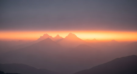 A majestic landscape view of mountain peaks silhouetted against a vibrant orange and purple gradient sky at sunset. Layers of mist and fog create a dramatic and atmospheric depth to the scene.の素材