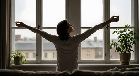A silhouette of a woman stretching her arms wide open in front of a window on a rainy day. The cozy indoor scene contrasts with the overcast weather outside, evoking feelings of waking up, relief, or freedom. A potted plant sits on the windowsill, adding a touch of nature to the peaceful morning atmosphere.の素材