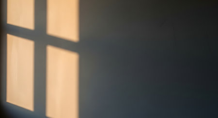The shadow of a window frame is cast sharply against a smooth, neutral-colored wall, illuminated by warm sunlight. The geometric lines create a minimalist and abstract composition, evoking a sense of calm and solitude. The interplay of light and dark suggests a quiet afternoon atmosphere.の素材