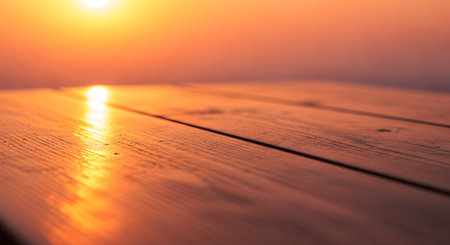 A close-up, low-angle shot of a wooden table surface bathed in the warm, golden light of a sunset. The background is softly blurred into a bokeh, creating a perfect copy space or backdrop for product placement.の素材