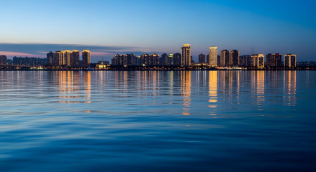 A panoramic view of a modern city skyline illuminated at night, reflected in the calm waters of a river or bay. The tall residential and commercial buildings glow with warm lights against a deep blue evening sky.の素材