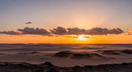 A breathtaking panoramic view of a vast desert landscape at sunset, featuring rolling sand dunes and rocky formations. The sun breaks through dramatic clouds, casting a golden glow over the arid wilderness.の素材