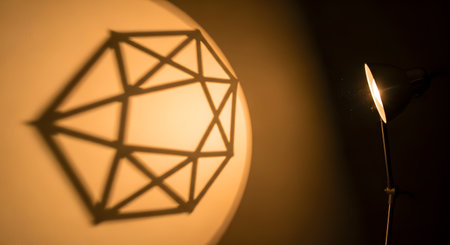 A desk lamp shines light onto a wall, casting a sharp, geometric shadow of a complex polyhedron structure. The image explores the interplay of light, shadow, and shapes in a minimalist and abstract composition.の素材