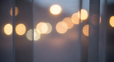 Blurred golden city lights create a beautiful bokeh effect seen through the texture of a sheer white curtain. The abstract composition suggests a cozy evening atmosphere inside looking out at the night.の素材