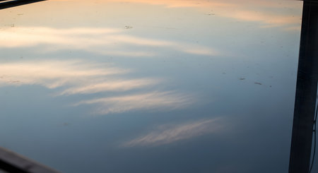 An abstract reflection of a sunset sky with streaky clouds on a smooth, reflective surface like water or glass. The image features soft hues of blue, white, and faint orange, creating a dreamy and atmospheric texture.の素材