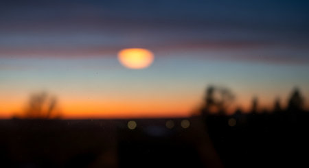 An abstract, blurred view of a sunset sky featuring warm orange and cool blue tones. The scene includes bokeh effects from city lights viewed through a window, creating a dreamy and atmospheric mood.の素材