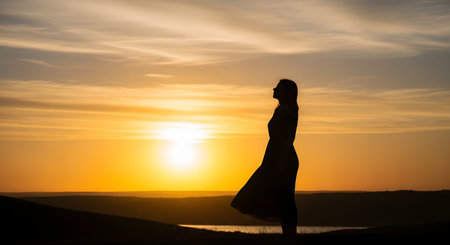 The silhouette of a woman in a dress standing against a vibrant golden sunset sky. The profile view captures a moment of peace, freedom, and connection with nature in a beautiful landscape.の素材