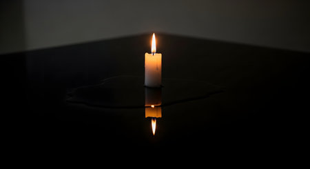 A single white candle burns brightly with a steady flame on a dark, reflective surface. The minimalist composition highlights the warm glow and the puddle of melted wax against the black background.の素材