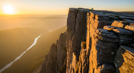 A breathtaking view of a rugged cliff edge illuminated by the golden light of sunrise, overlooking a winding river in a forest valley. The dramatic landscape features towering rock formations and a hazy, atmospheric horizon.の素材