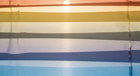 Strips of colorful fabric in shades of orange, yellow, white, and blue are backlit by the sun, creating a vibrant and translucent texture. The horizontal lines and soft light make for a beautiful abstract background.の素材