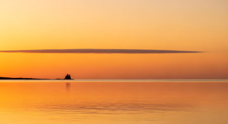 A serene and minimalist seascape featuring a golden sunset with a perfectly calm ocean reflecting the orange sky. A small, distant island silhouette sits on the horizon under a thin strip of cloud, creating a peaceful atmosphere.の素材