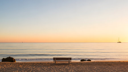 An empty wooden bench rests on a wide, sandy beach facing a tranquil ocean at sunset or sunrise. The sky is a gradient of soft orange and blue, with the horizon line emphasized, creating a minimalist and serene scene with small boats in the distance.の素材
