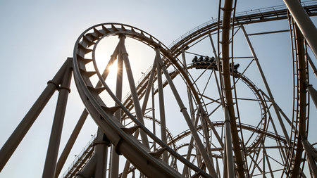 A dramatic low-angle view of a complex steel roller coaster track featuring a large loop and twisting rails. The sun bursts through the structure as a coaster train blurs by, capturing the excitement of an amusement park ride.の素材