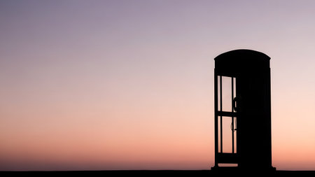 The silhouette of a classic telephone booth stands out against a beautiful gradient sunset sky fading from purple to orange. The image evokes a sense of nostalgia and communication in a serene evening setting.の素材