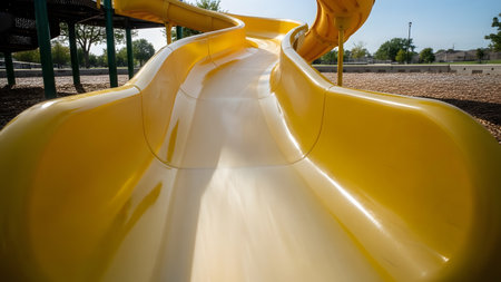 A point-of-view perspective looking down a bright yellow plastic slide at an outdoor playground. The curve of the slide leads the eye toward the gravel ground and park surroundings on a sunny day.の素材
