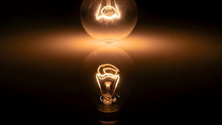 A close-up of a glowing tungsten filament inside a clear lightbulb, reflecting on a glossy surface against a dark background. The warm orange light symbolizes ideas, innovation, and energy.の素材