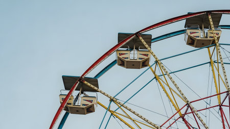 A low-angle view of colorful Ferris wheel gondolas against a clear blue sky. The red, yellow, and blue structure represents classic amusement park fun and summer entertainment.の素材