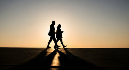The silhouette of a man and a woman walking side-by-side against a bright, golden sunset sky. The minimalist composition focuses on their shadows and the warm light, suggesting companionship, travel, or a journey.の素材