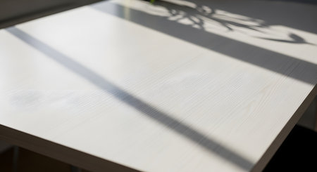 A close-up view of a light-colored wooden table surface with soft, abstract leaf shadows cast by sunlight. The image serves as a clean, natural background with texture and copy space.の素材