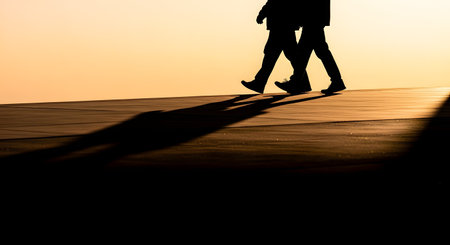 The silhouettes of two people walking side-by-side on a flat surface against a golden sunset background. Long, dramatic shadows stretch out before them, suggesting companionship, a journey, or an evening stroll.の素材