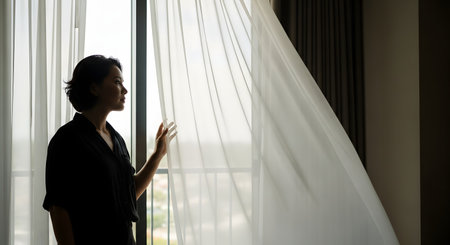 A woman stands in profile by a large window, gently holding back a sheer white curtain to look outside. The soft daylight illuminates her face, creating a mood of quiet contemplation, waiting, or enjoying the view.の素材