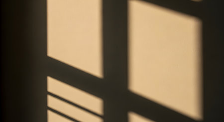Abstract geometric shadows cast by a window frame onto a smooth beige wall. The sharp contrast between the warm light and the dark lines creates a minimalist and artistic composition, evoking a sense of late afternoon sunlight.の素材