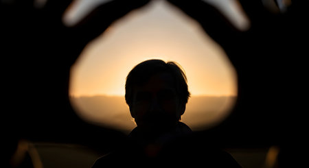 A silhouette of a person is creatively framed by two hands forming a shape against a warm, golden sunset background. The image evokes feelings of connection, perspective, love, and artistic vision in a natural setting.の素材