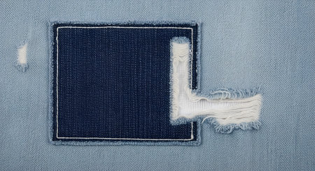 A detailed close-up of blue denim fabric featuring a dark blue rectangular patch with white frayed edges and contrast stitching. The image highlights textile texture, clothing repair, and fashion design details.の素材