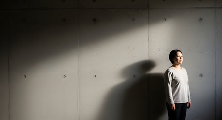 A woman stands stoically against a textured concrete wall, illuminated by a beam of light that casts a dramatic shadow. The minimalist composition and high contrast lighting create a mood of strength, solitude, or contemplation.の素材
