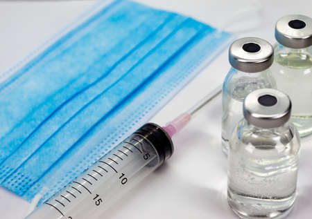 Coronavirus COVID-19 Vaccine Vial, Syringe and blue Medical Face Mask on white background - close up...