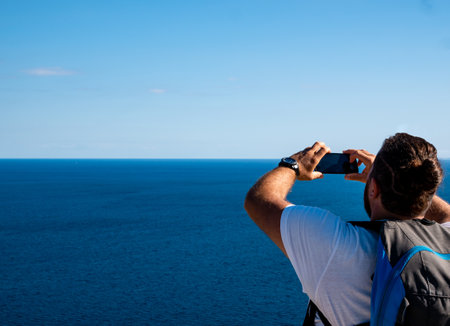 Young caucasian man takes a picture with his smartphone at the sea and the horizon on a summer dayの写真素材