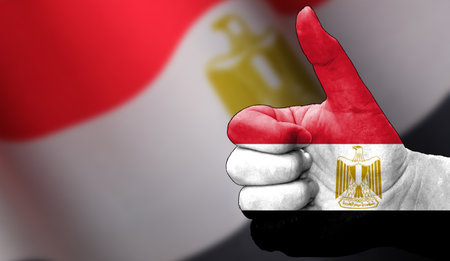 hand with thumbs up in approval with the Egyptian flag painted. Image with flag background area out of focus, copy space areaの写真素材