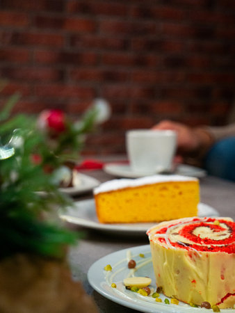 Close-up of two slices of decorated cake on plates, with a white cup of coffee and blurred festive...