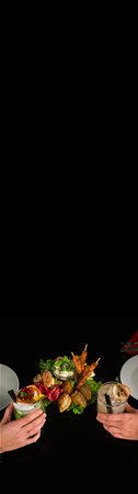 Overhead shot of two hands holding cocktails on a black table. A plate of mixed appetizers is in the...