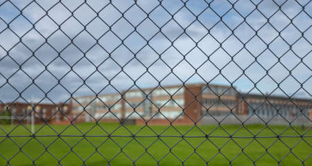 A Typical High School Building And Sports Field Under A Cloudy Sky With Focus On The Security Fence...